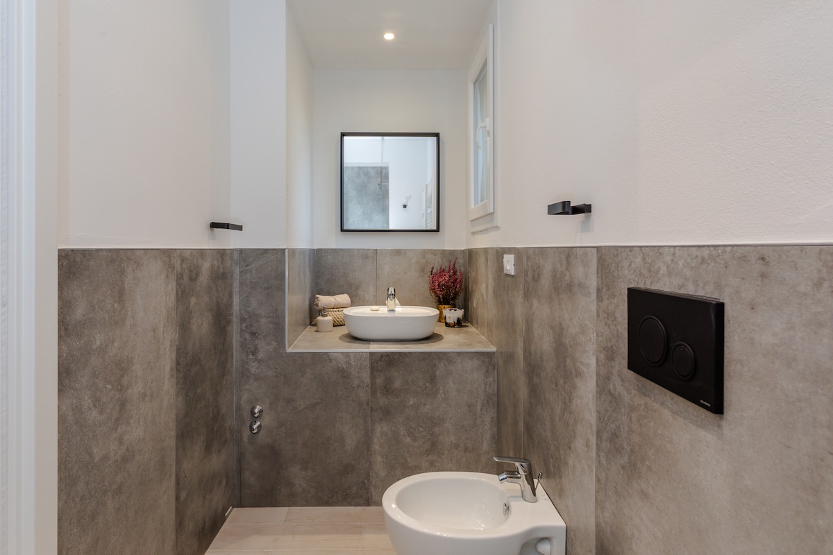A modern bathroom is displayed with a minimalist design. A large mirror reflects the space, while a stylish sink is positioned on a gray stone counter. Light-colored tiles cover the floor, and a small decorative plant adds a touch of nature.