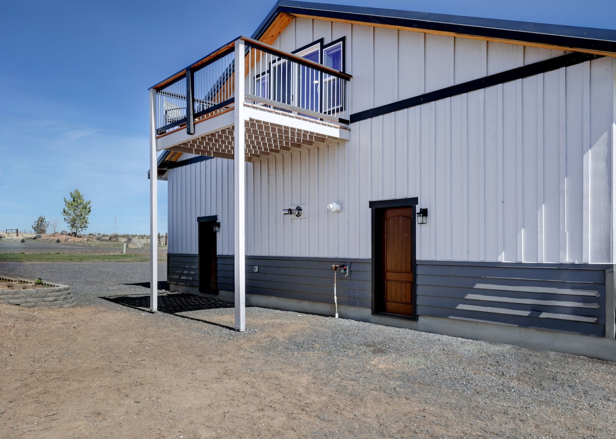 The exterior of the studio showcases a two-story structure with a raised balcony above a covered entryway. The building features a combination of white paneling and dark grey accents, with windows and a door framed in matching hues, set against a backdrop of open land.