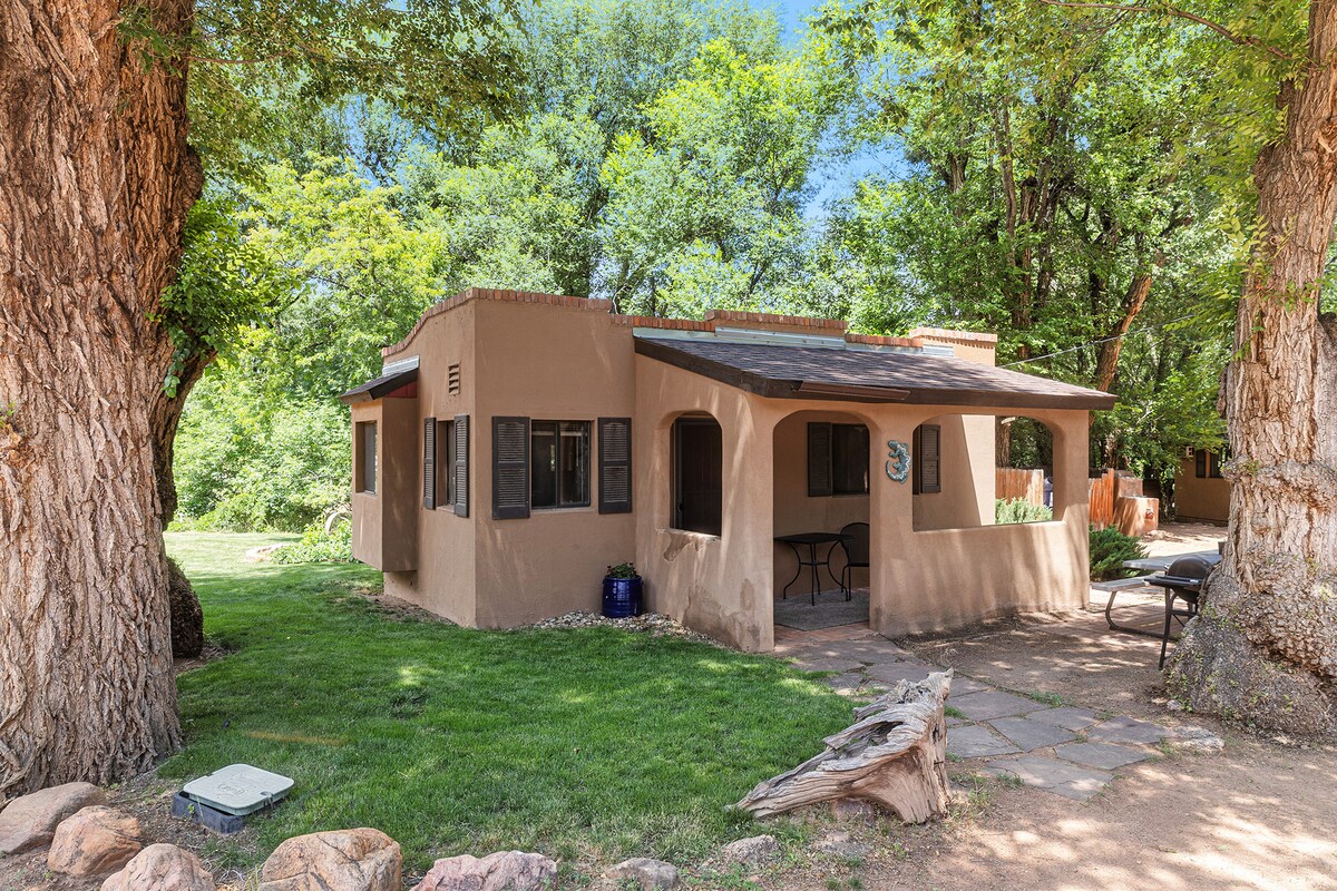 An adobe cottage is nestled within lush greenery, framed by large trees. A covered porch features comfortable seating, while large windows provide natural light. The well-kept lawn contributes to a serene outdoor space, complete with a nearby BBQ grill.