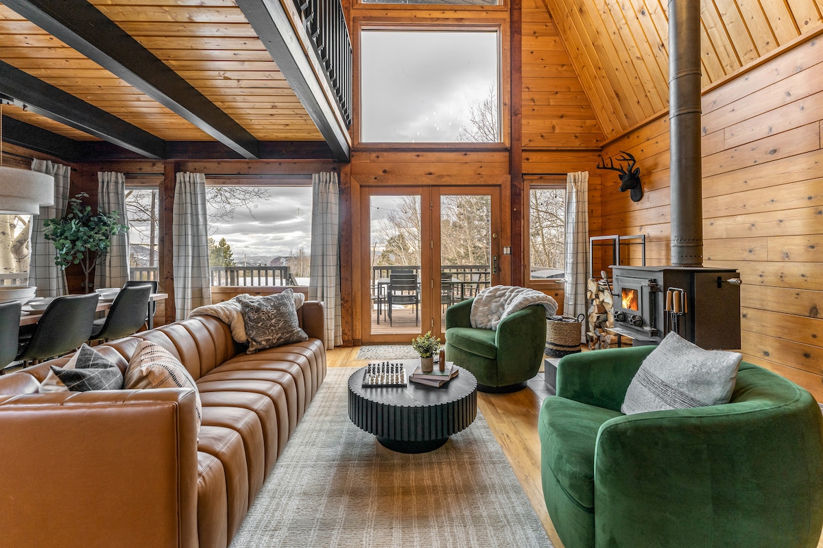 A spacious living area features a leather sectional sofa and two green chairs, positioned around a round coffee table. Large windows offer natural light, while a wood-burning stove provides warmth against the wooden paneling and vaulted ceiling.