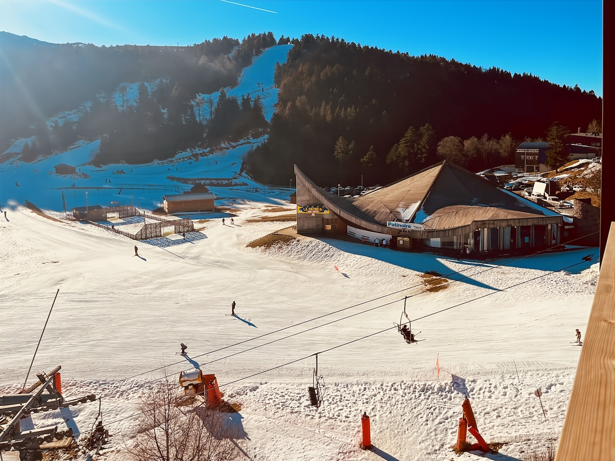 A snow-covered landscape is depicted, featuring a ski lift and skiers navigating the slopes. A uniquely designed building with a sloped roof is visible, surrounded by snow-capped mountains and evergreen trees. Bright sunlight illuminates the scene, enhancing the wintry atmosphere.