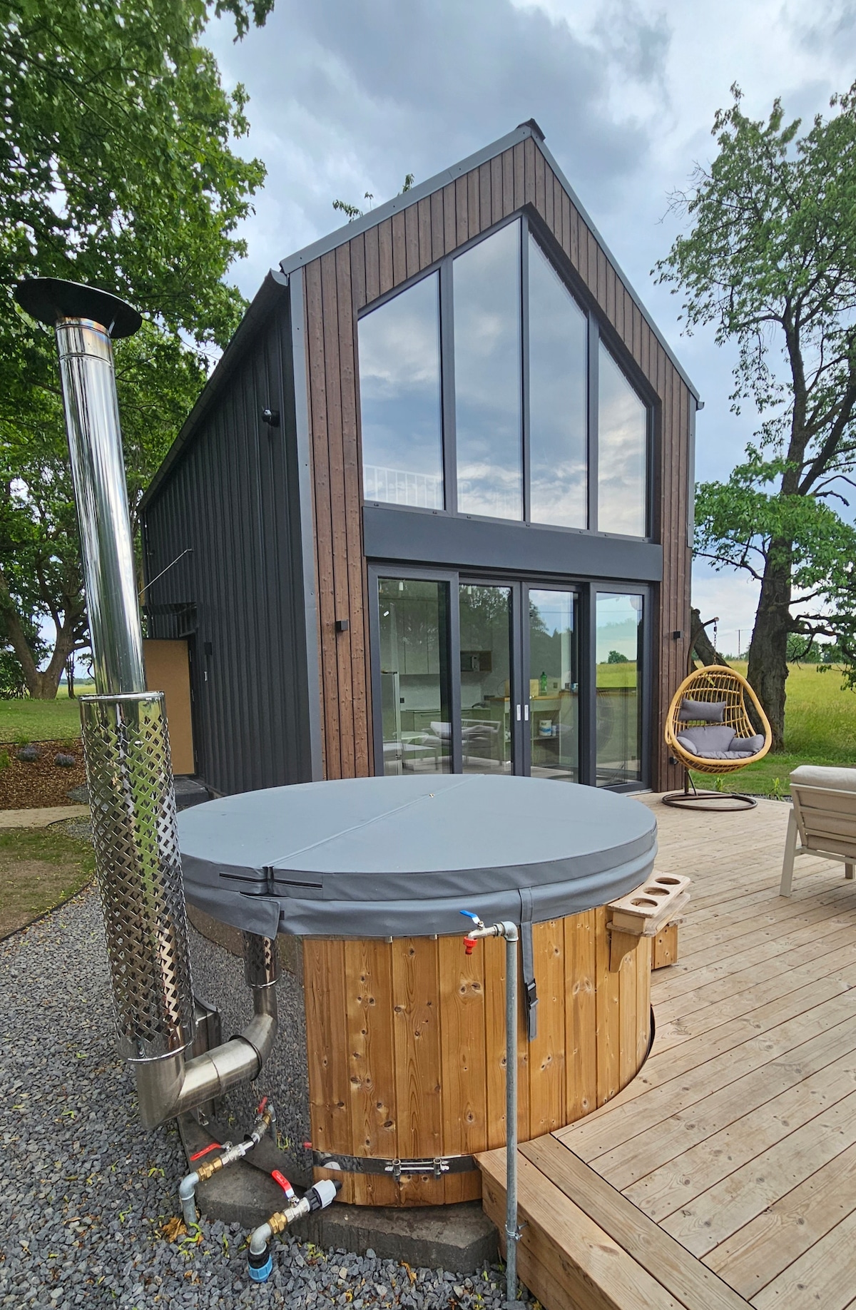 A modern cabin exterior is shown, featuring large glass windows that fill the interior with natural light. A wooden hot tub is positioned on a wooden deck, surrounded by gravel. A stylish hanging chair can be seen nearby, framed by greenery.
