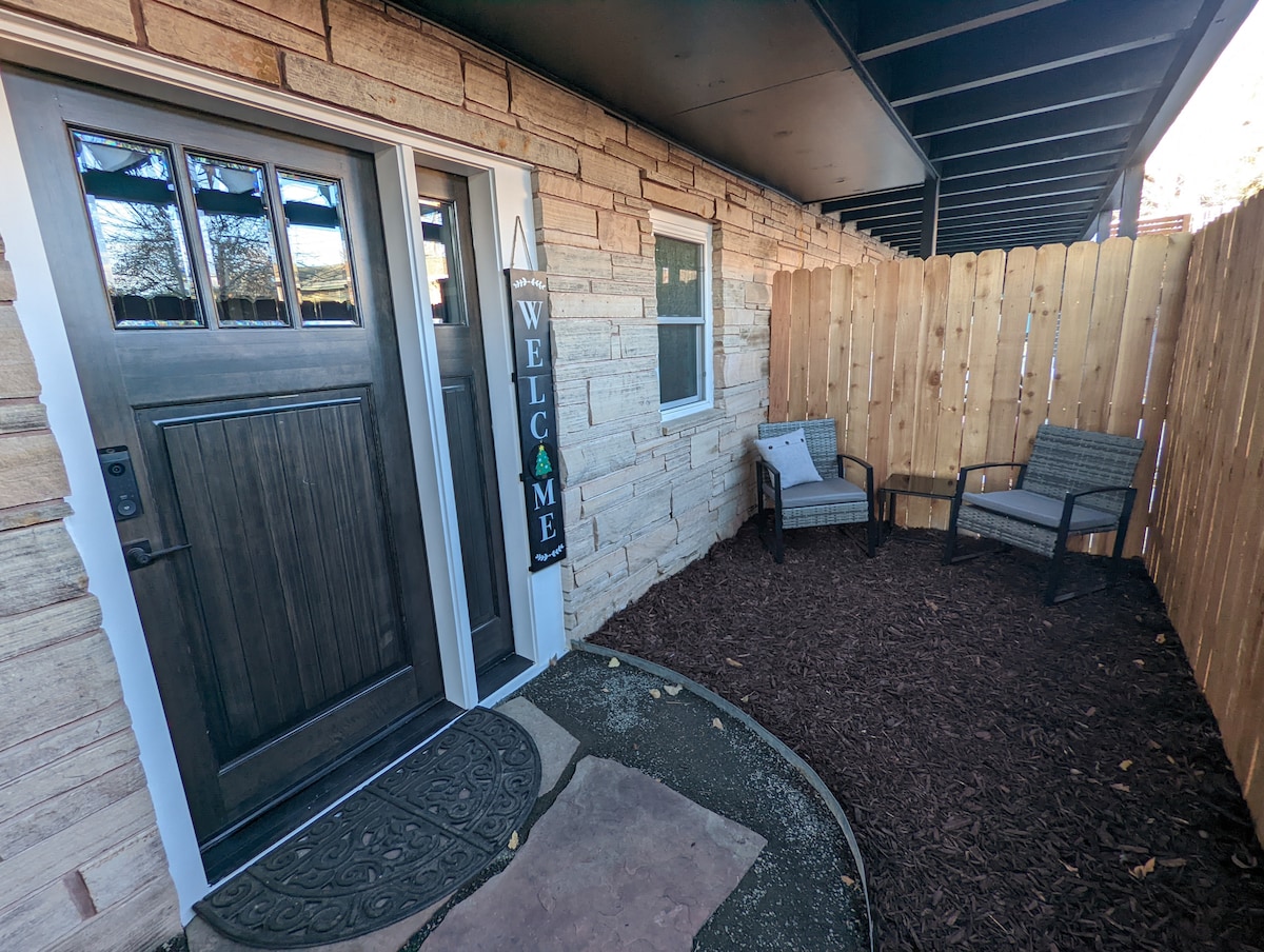 A welcoming entrance is highlighted by a wooden door with glass panels. A decorative 'Welcome' sign hangs nearby. Two outdoor chairs are positioned on mulch, surrounded by a fenced area providing privacy.