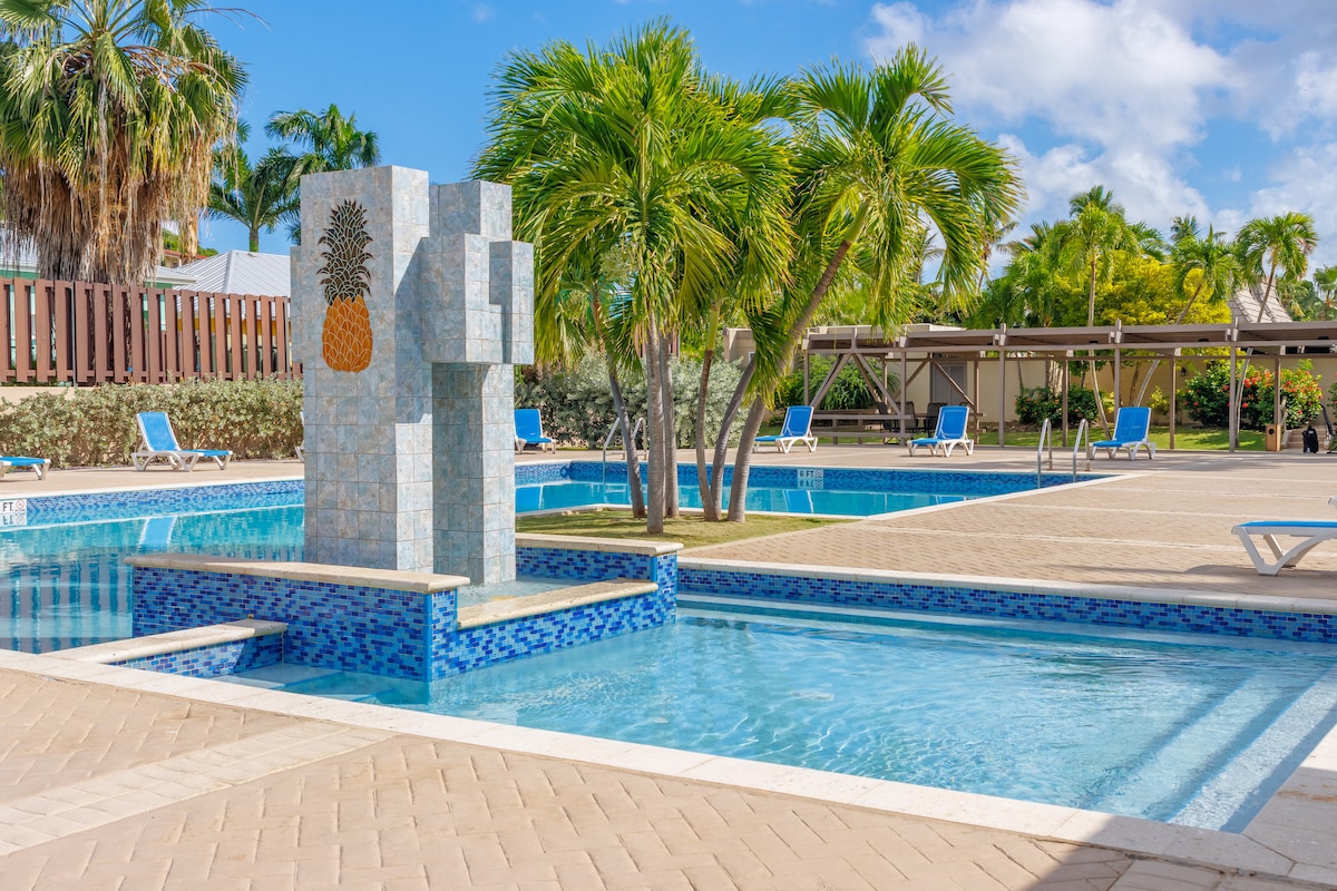 A community pool area features a central fountain adorned with a pineapple motif, surrounded by palm trees. Lounge chairs are placed along the pool's edge on tiled decking, with clear blue water reflecting the sky and nearby greenery.