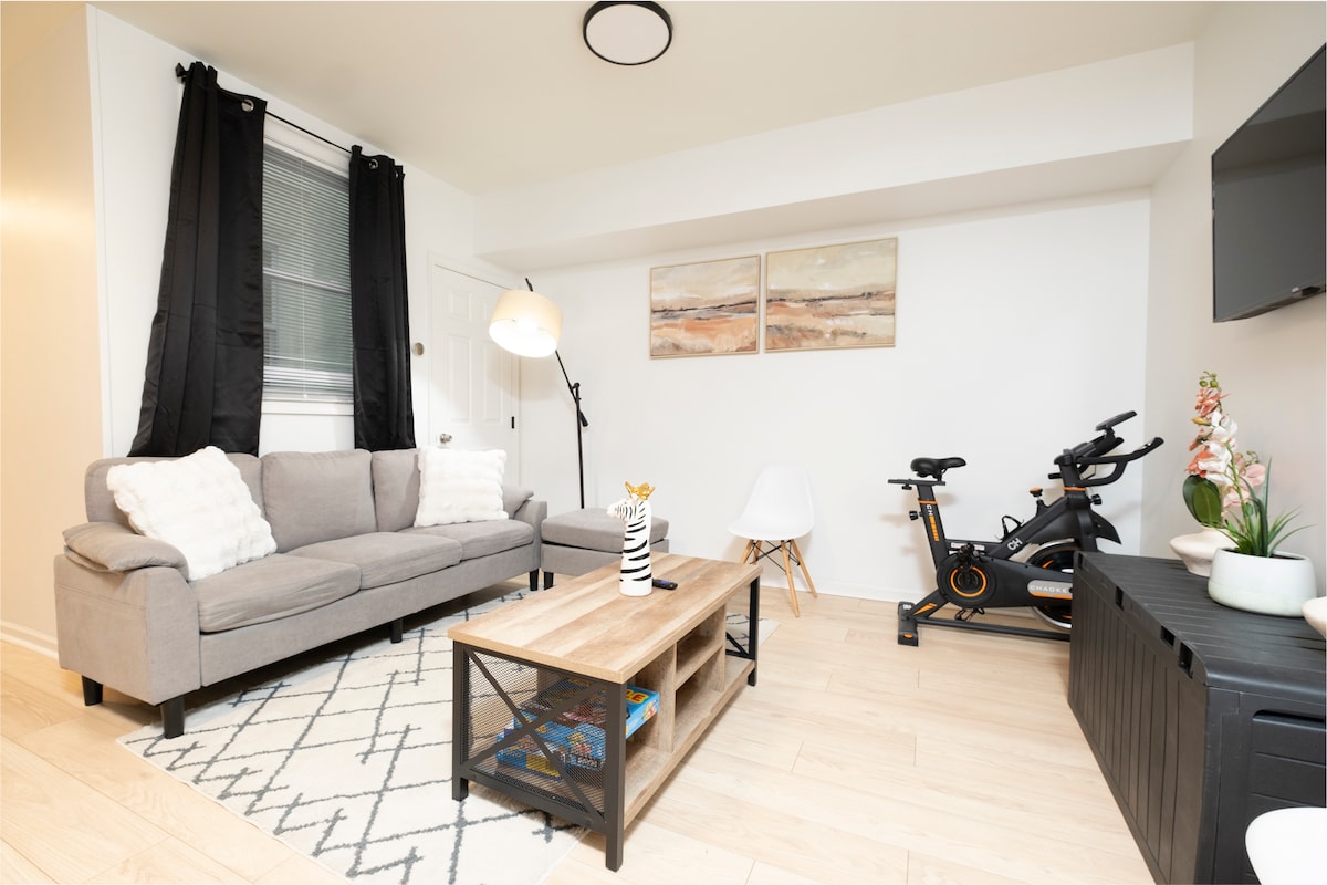 A living area features a gray sofa adorned with decorative pillows, positioned in front of a wooden coffee table. A fitness bike is located to the right, alongside a small potted plant. Black curtains frame the window, and abstract artwork is displayed on the wall.