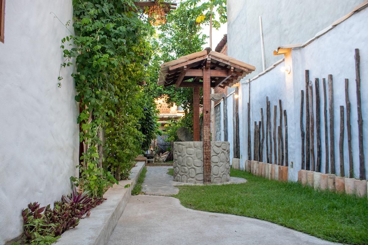 A pathway lined with greenery leads to a rustic well, surrounded by walls featuring natural textures. A grassy area to the side adds a touch of nature, while slender wooden posts enhance the outdoor setting.
