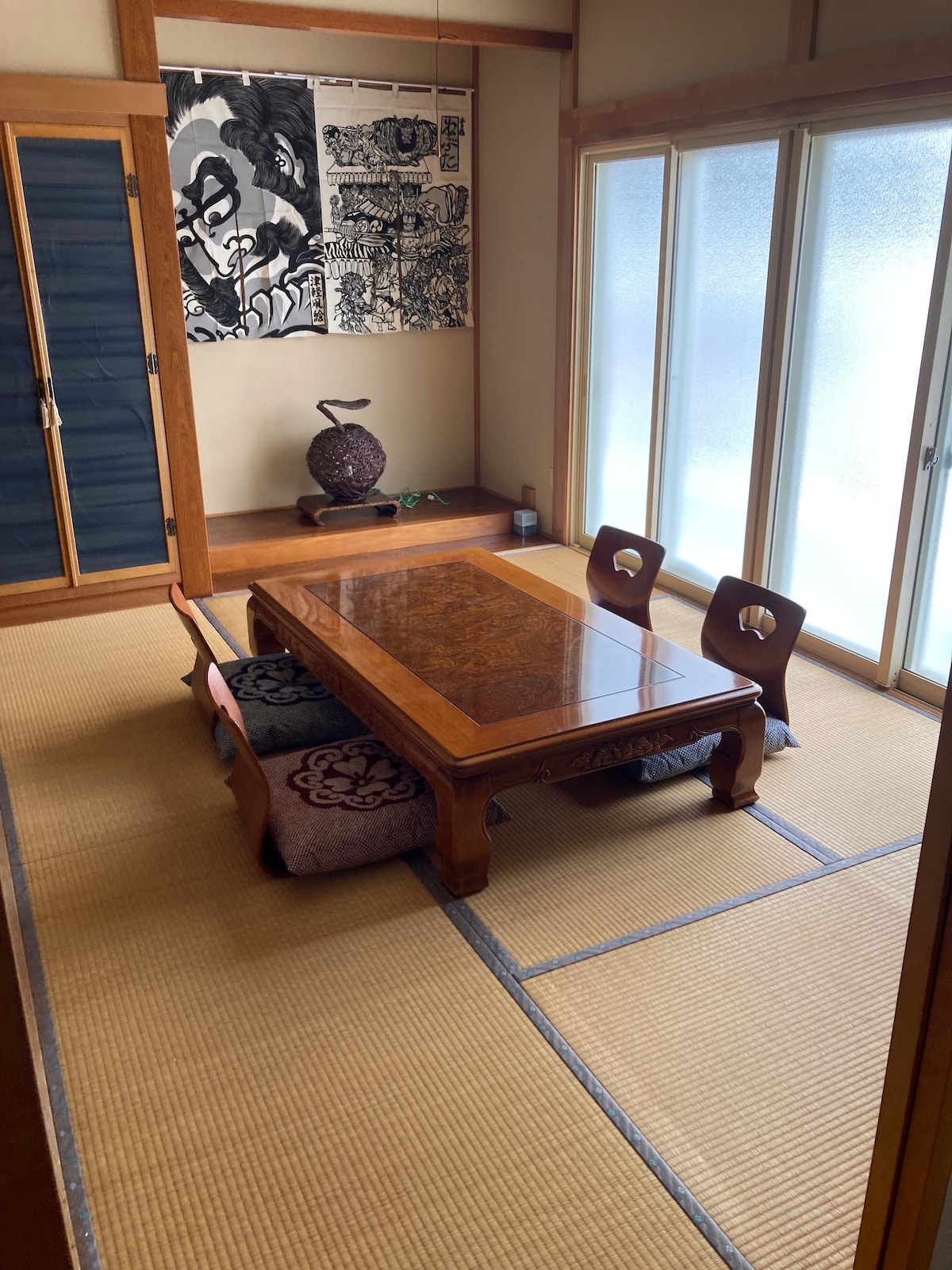 A traditional Japanese-style room features a wooden low table surrounded by two seating cushions. A decorative piece sits on the floor nearby, and sliding glass doors allow natural light to illuminate the space while providing views of the outside.