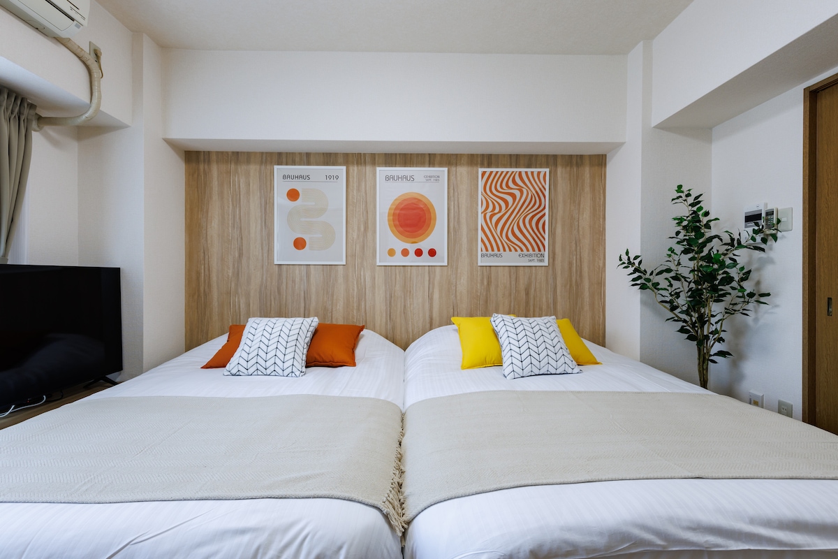 The sleeping area features two spacious beds with white linens and decorative pillows in yellow and gray. Above the beds, three art prints add a colorful touch. A decorative plant stands in the corner, enhancing the room’s comfort.