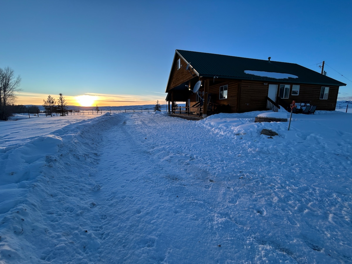A rustic cabin is situated in a snowy landscape, with a pathway leading towards the entrance. The sun sets in the background, casting a warm glow across the white snow. The cabin features a green roof and a covered porch, enhancing its mountain retreat charm.