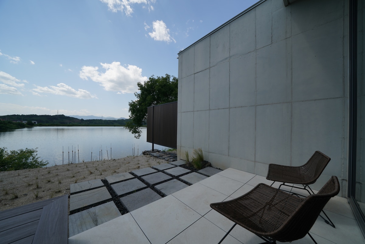 A tranquil outdoor terrace features two woven chairs placed on a tiled surface. The area overlooks a calm body of water, bordered by natural landscaping. A gentle slope leads to the water's edge, with a few trees visible in the background under a partly cloudy sky.