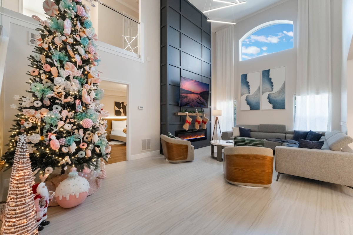 A spacious living area is highlighted by a beautifully decorated 12-foot Christmas tree and soft seating arrangements, including a large sectional and two wooden chairs. Natural light streams in through large windows, illuminating the modern decor and enhancing the cozy ambiance.