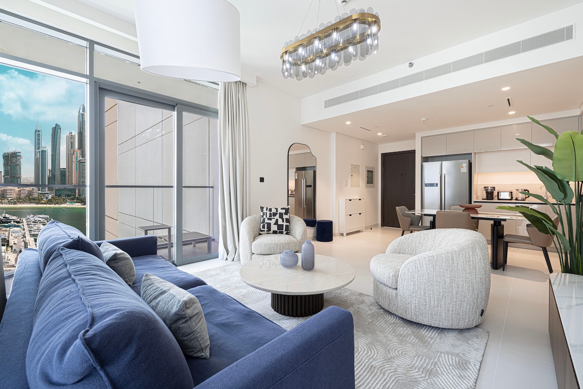 A modern living area showcases a plush blue sofa and two circular armchairs, set around a round coffee table. Large glass doors open to a balcony with views of Dubai's skyline. Soft natural light fills the space, complemented by a contemporary chandelier above.