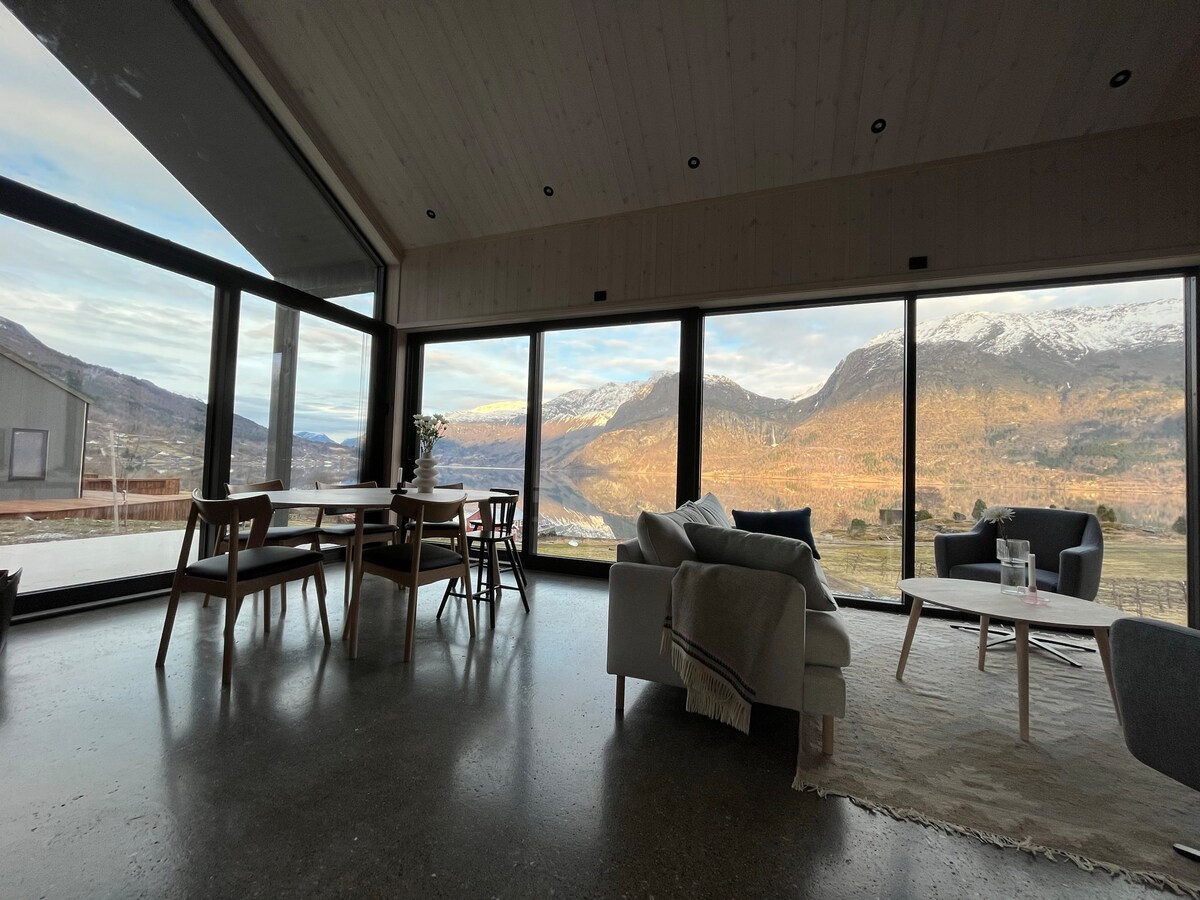 The open-plan living area features large panoramic windows offering breathtaking views of the surrounding mountains and landscape. A modern dining table is set for four, paired with wooden chairs. A cozy seating arrangement is positioned near the window, complemented by a soft area rug.
