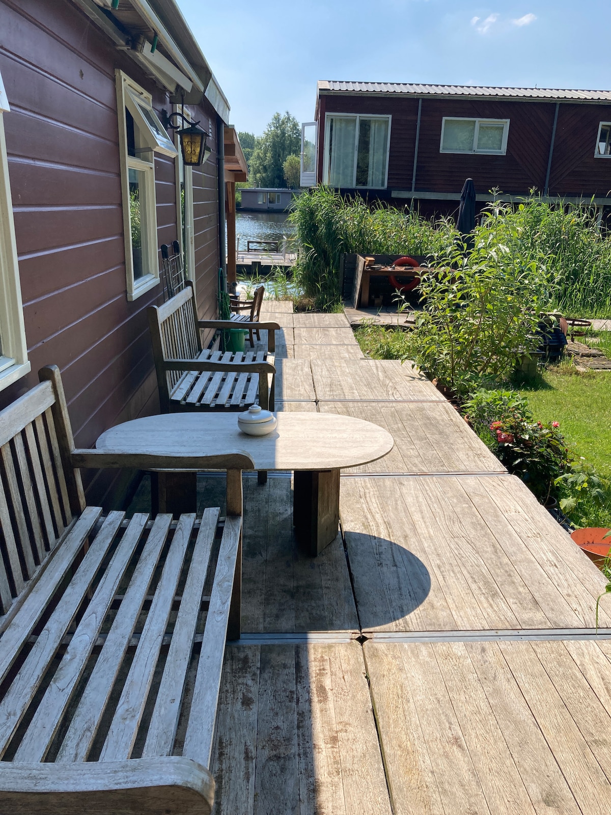 A wooden terrace is shown with two benches and a circular table, positioned along a pathway. Lush greenery lines the sides, and the calming water view is visible in the background, creating a serene outdoor seating area.