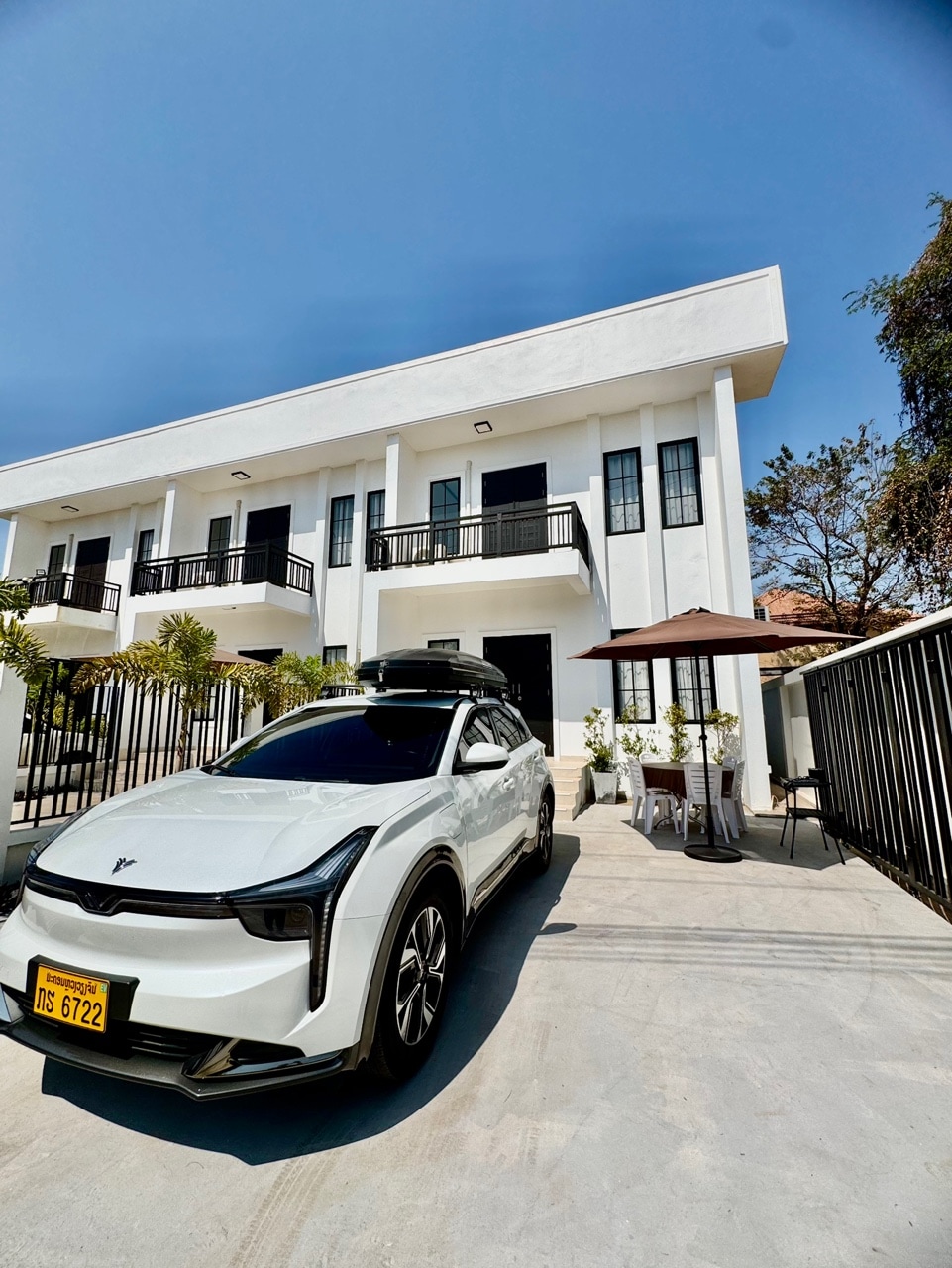 An exterior view of a modern white building with spacious balconies and railings. A sleek vehicle is parked in the driveway, under a bright blue sky. Green plants are positioned near the entrance, and an umbrella-shaded seating area is visible to the side.