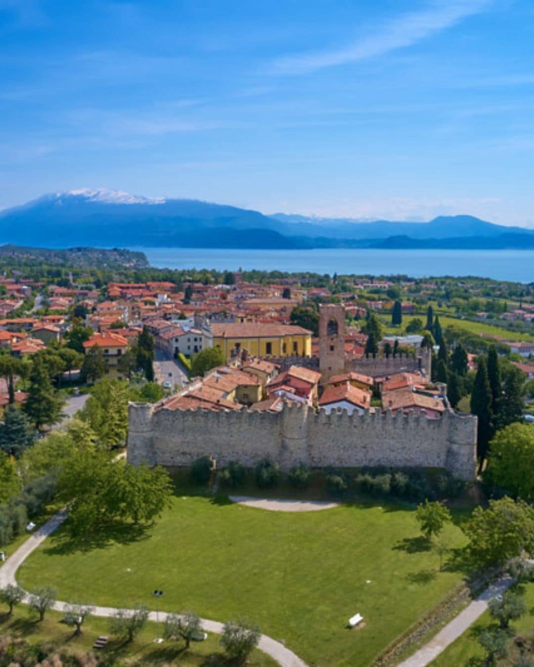 An aerial view captures a medieval castle surrounded by well-preserved walls, set against a backdrop of rolling hills and the shimmering Lake Garda. A quaint village with traditional architecture is visible beside the castle, and lush greenery enhances the peaceful landscape.