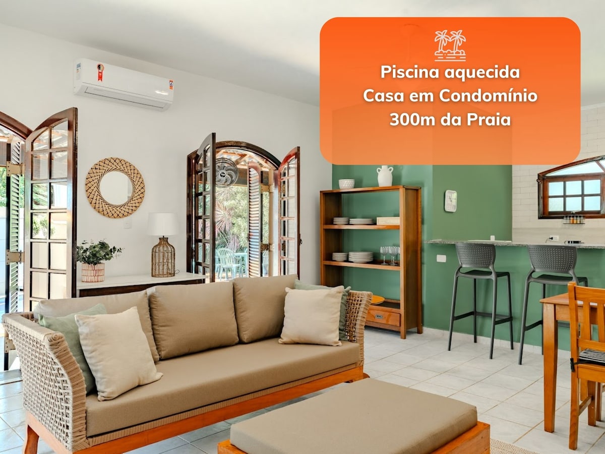 A welcoming living area features a light-colored sofa accompanied by wooden chairs and a dining table. The space is organized with an open kitchen and bar stools, and large windows allow natural light to fill the room.