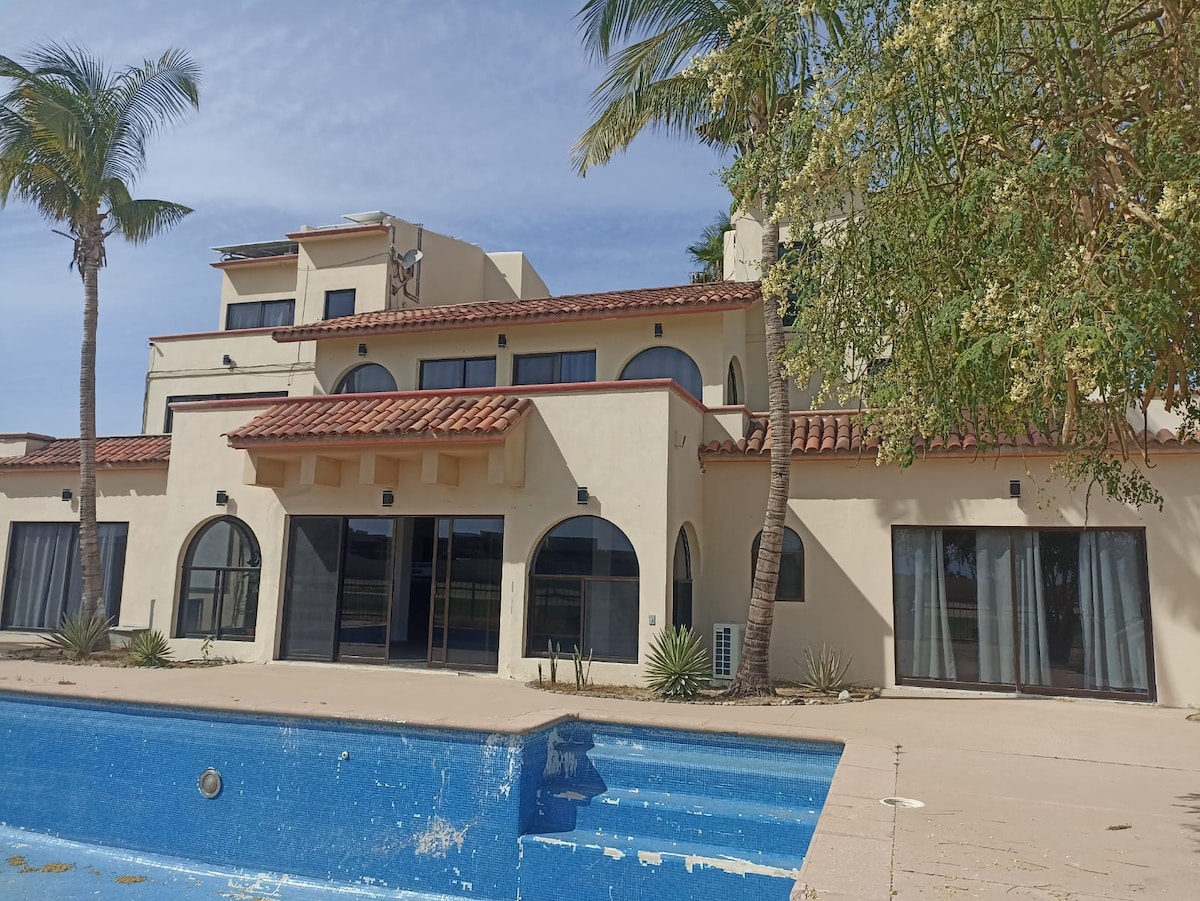 The exterior of a multi-level villa is displayed, featuring arched windows and a tiled roof. Palm trees are positioned nearby, and a swimming pool with a faded blue surface is partially visible in the foreground, enhancing the inviting atmosphere of the property.