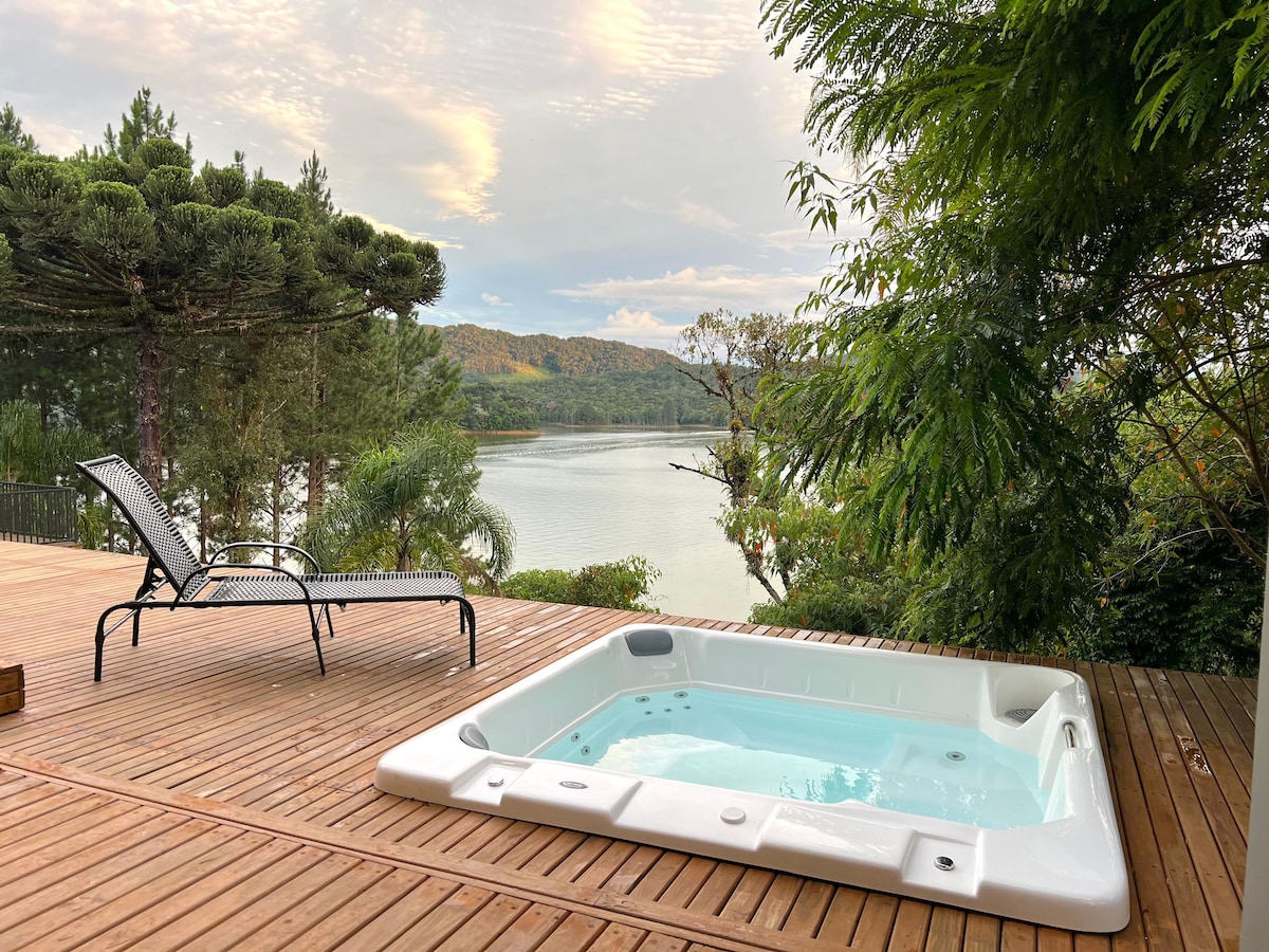 A wooden deck features a spacious hot tub, inviting relaxation with water visible. A lounge chair is placed nearby, offering a comfortable spot to enjoy the surrounding nature and serene views of the river and lush greenery.