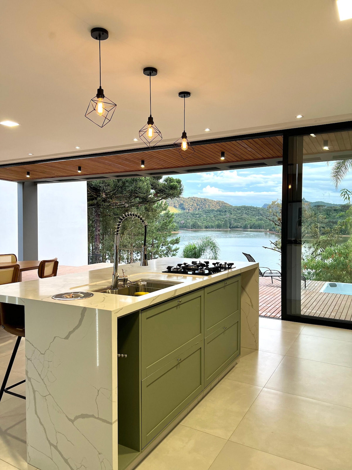 A modern kitchen is showcased, featuring a sleek green island with a gas cooktop and a stylish sink. Large sliding glass doors offer a view of the serene water and surrounding nature, while industrial-style light fixtures hang above the space.