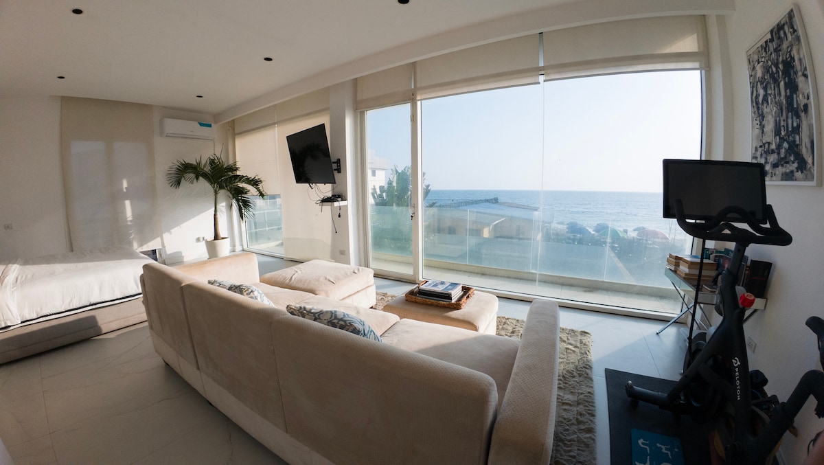 A bright living area is fitted with a large, comfortable sofa that faces expansive windows. Natural light floods the space, offering views of the Atlantic Ocean. A small indoor plant adds a touch of greenery, while a stationary bike is positioned nearby.