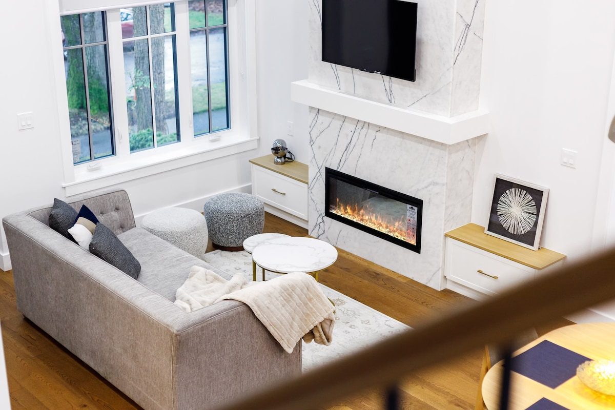 A modern living area features a grey sectional sofa positioned near a sleek marble fireplace. A round coffee table centers the space, accompanied by an ottoman. Large windows allow natural light, while a television is mounted above the fireplace, enhancing the cozy ambiance.