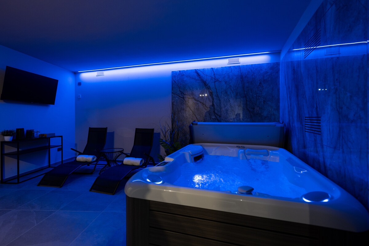 An exclusive wellness area hosts a heated hot tub with adjustable hydro-massage features, illuminated by soft blue lighting. Two reclining loungers are positioned nearby, creating a serene environment for relaxation. A wall-mounted television is visible, complementing the modern design.