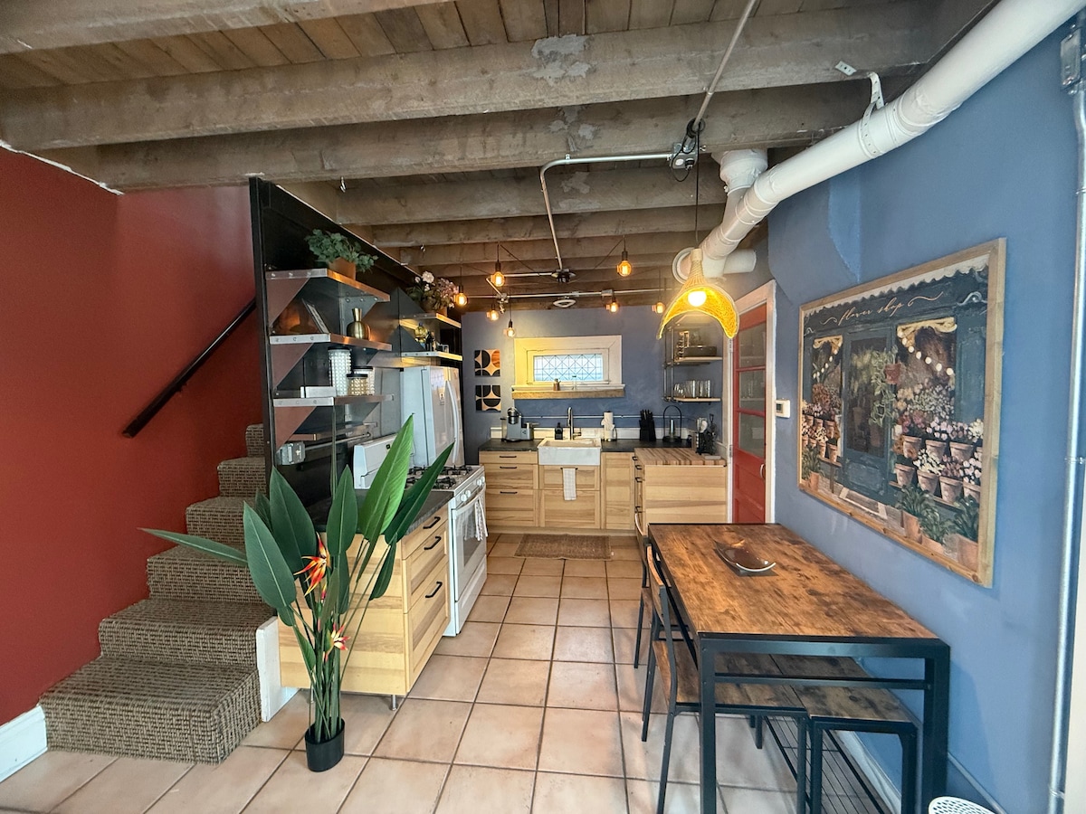 The kitchen area features modern appliances integrated into wood cabinetry. A ceramic stove and oven are complemented by a large sink. A dining table with dark metal legs offers seating, while decorative plants add a touch of greenery. Warm light fixtures illuminate the space.