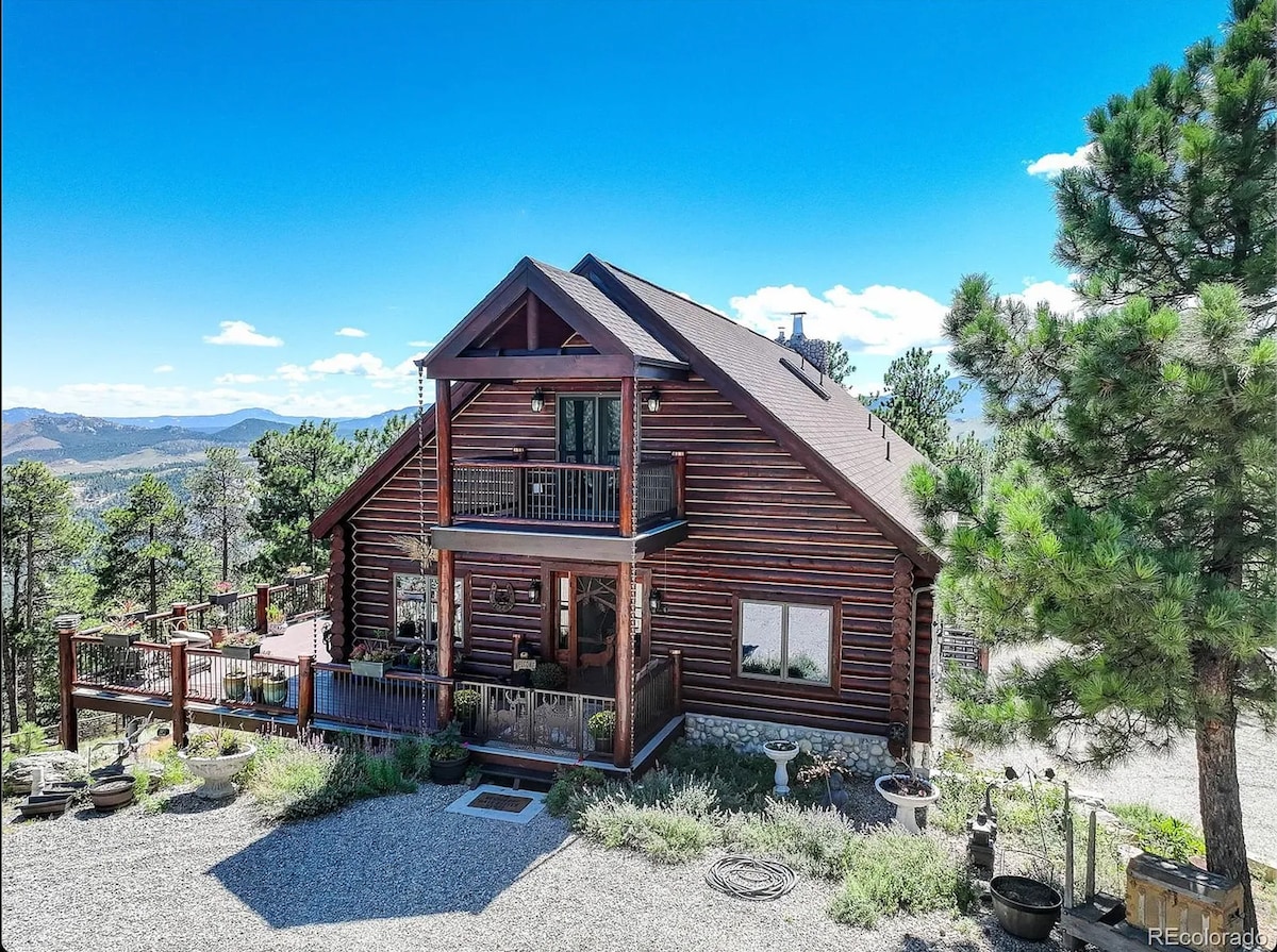 A spacious log home is situated among tall pine trees, featuring a wrap-around deck that offers views of the surrounding landscape. Large windows allow natural light to fill the interior. The exterior includes a gravel path leading to the entrance surrounded by garden elements.