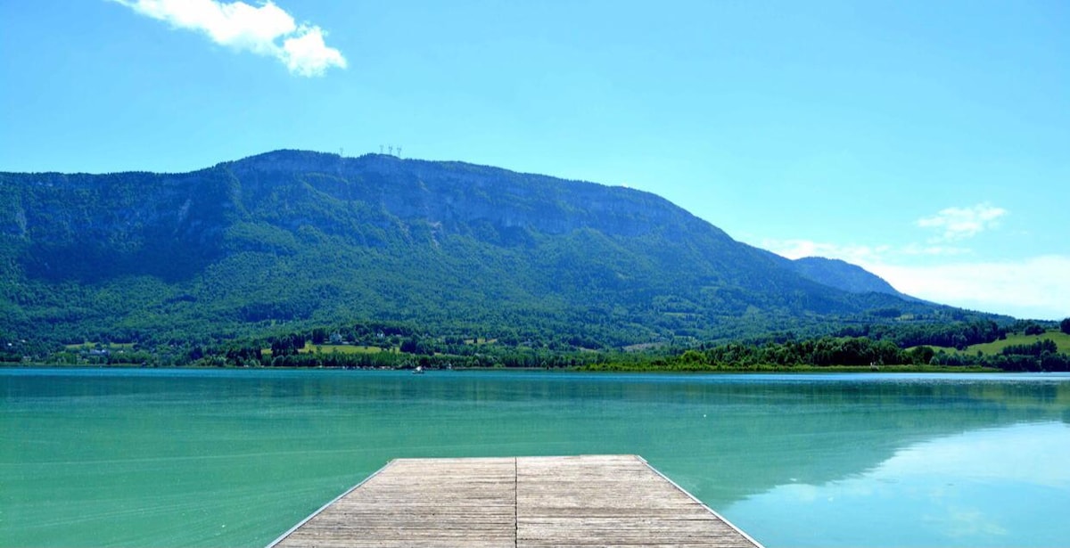 A wooden dock extends into a calm, clear lake, surrounded by lush green mountains. The scene is illuminated by bright sunlight, creating a serene atmosphere with gentle ripples on the water's surface.
