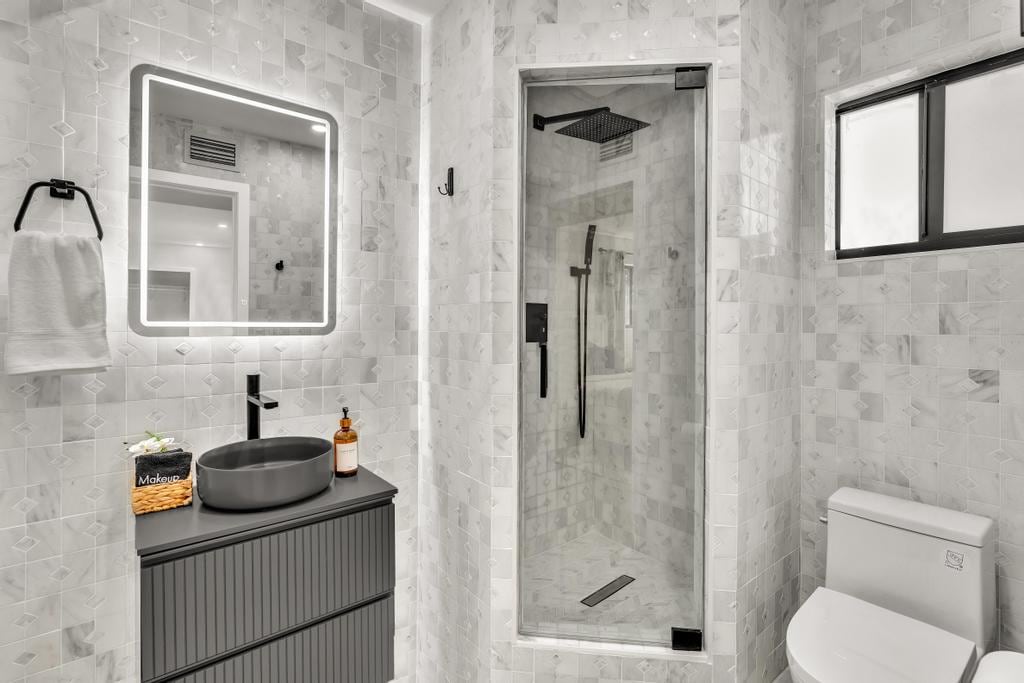 A modern bathroom featuring white marble tiles and sleek fixtures. A spacious shower with a glass enclosure is prominently displayed. The dark vanity contrasts with the light tiles, equipped with a round sink and organized toiletries. Soft lighting is provided by a backlit mirror.