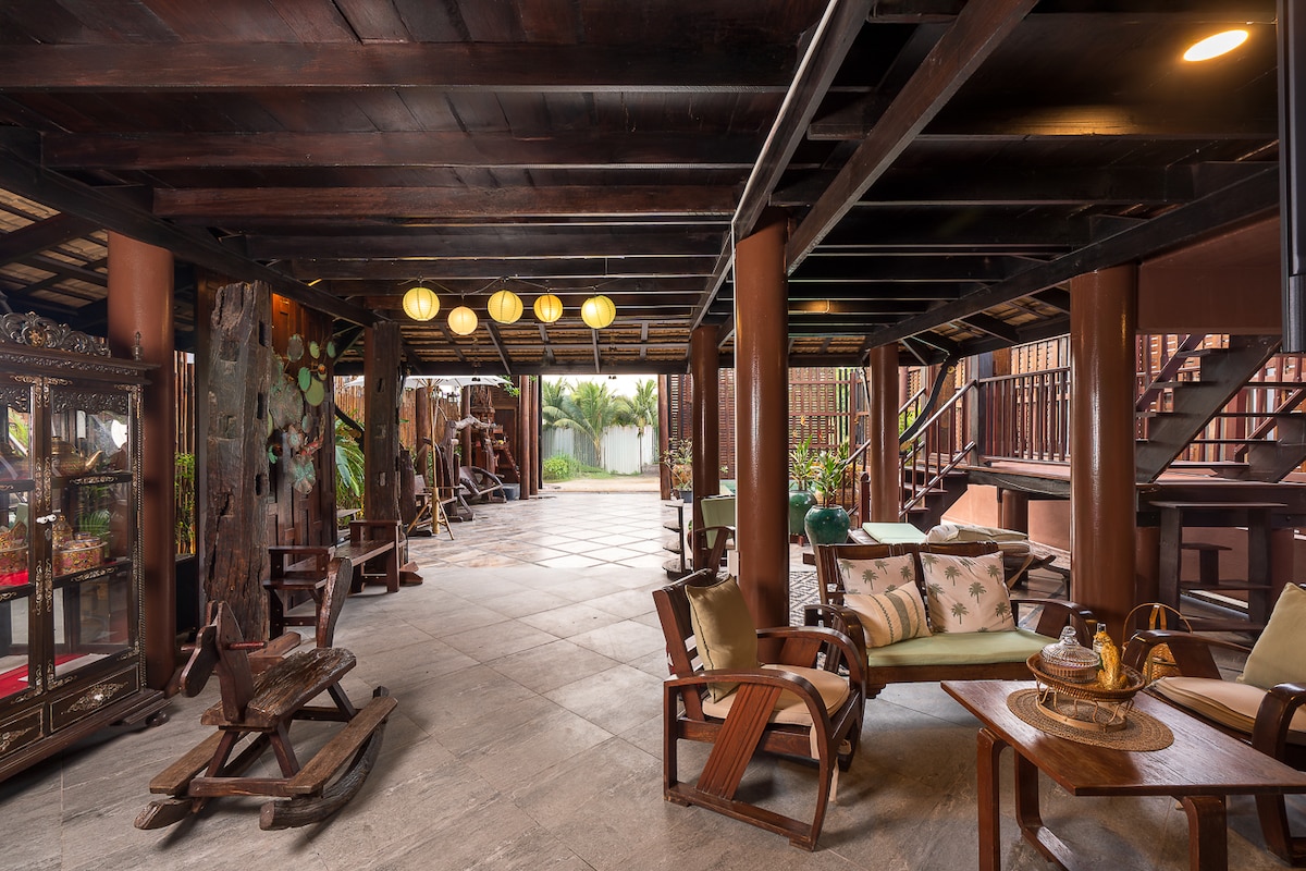 A spacious open-air living area features wooden beams and a stone floor. Elegant lanterns hang from the ceiling, and traditional wooden furniture is arranged for comfort. Lush greenery is visible through large openings, enhancing the connection to nature.