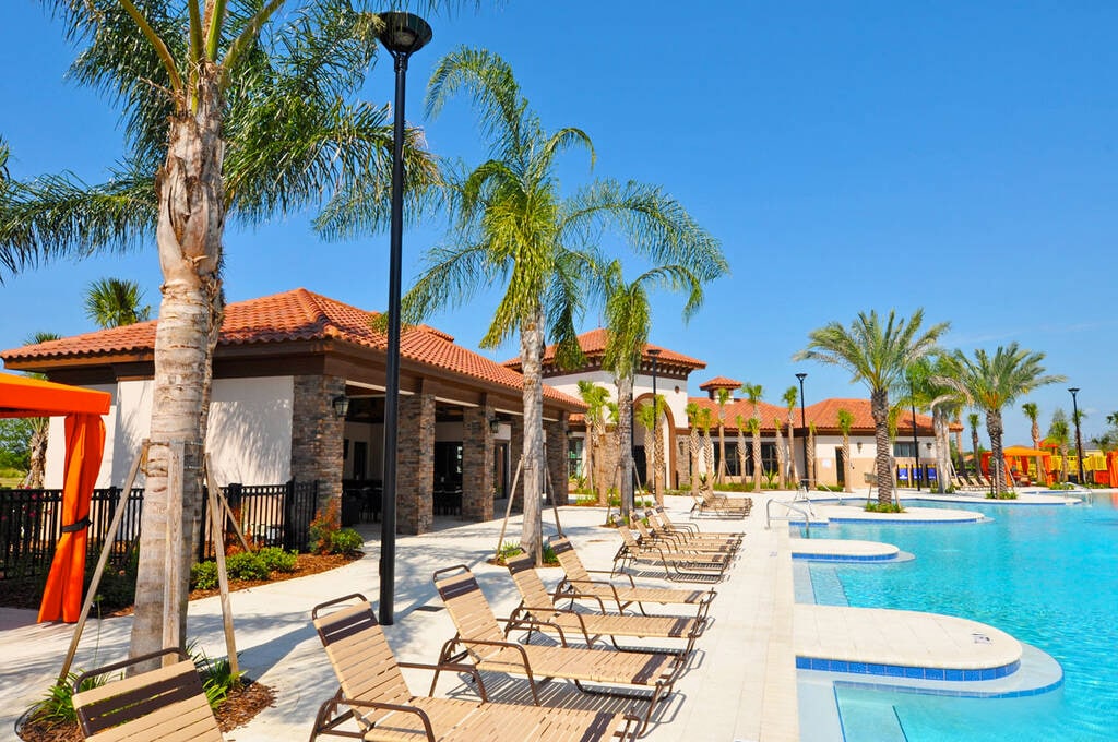 A bright resort area is depicted, featuring a large pool surrounded by sandy-toned loungers under palm trees. An inviting clubhouse with a brick exterior stands in the background, complemented by vibrant orange cabanas adding a tropical touch to the outdoor setting.