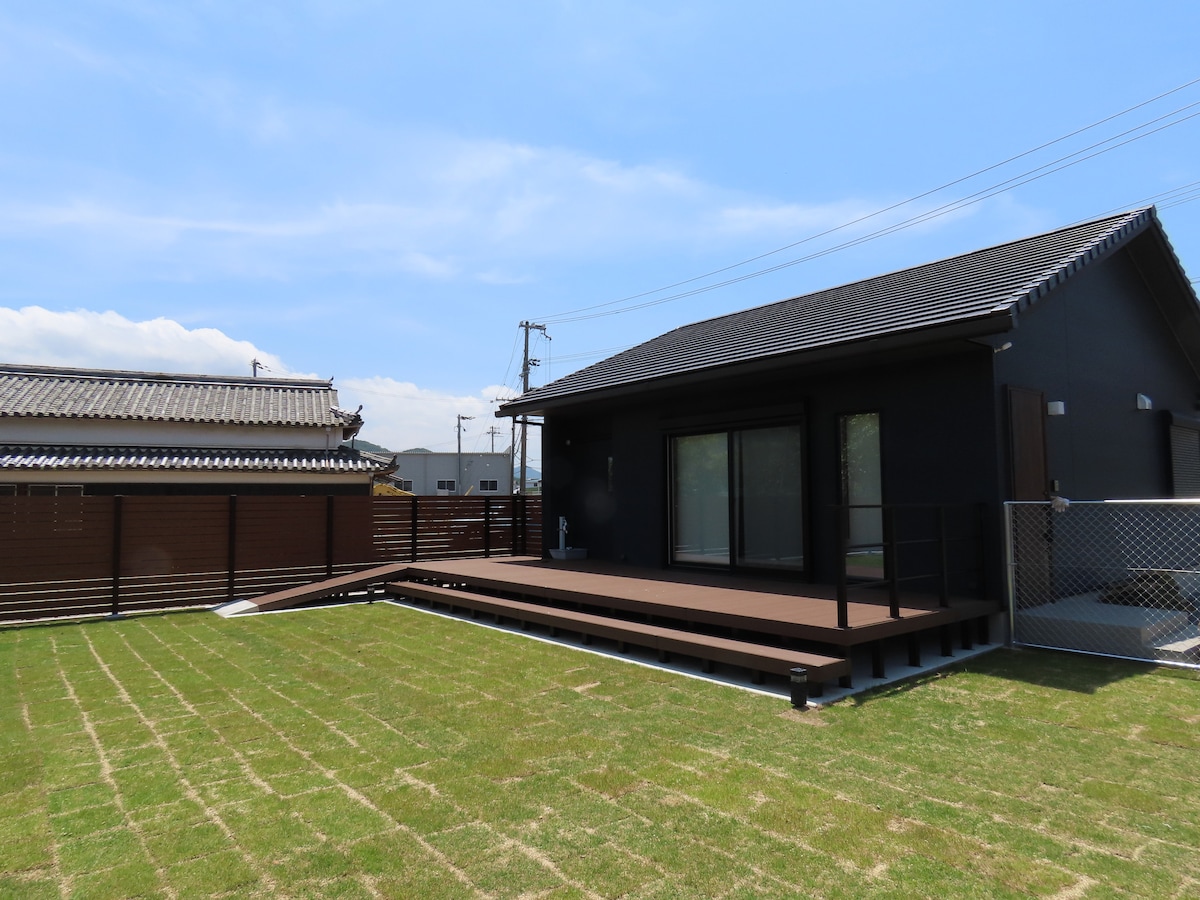A modern cottage is set against a clear blue sky, featuring a spacious wooden deck that extends towards a well-maintained grassy yard. The structure has large windows allowing for ample natural light, and fencing provides privacy around the outdoor area.