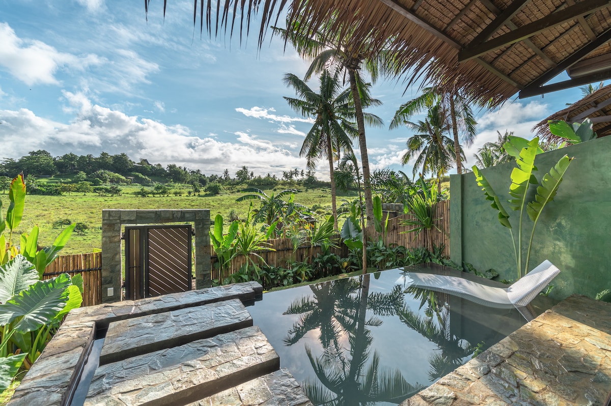 A private infinity pool is surrounded by tropical greenery, offering views of lush rice fields and palm trees. The serene water reflects the sky, while a sun lounger rests at one side. Natural stone elements enhance the tranquil atmosphere of the outdoor space.