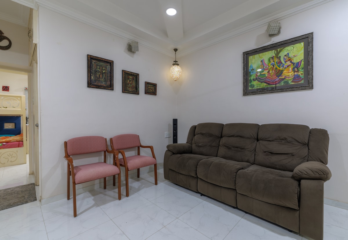 The living room features a comfortable brown sofa alongside two wooden-framed chairs with soft upholstery. Artwork adorns the walls, including a colorful framed piece. A decorative light fixture hangs from the ceiling, providing soft illumination.