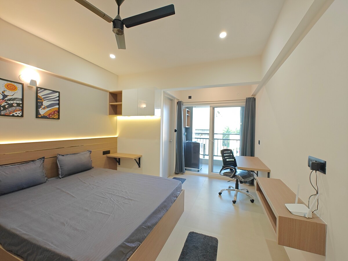The private bedroom features a spacious bed with neutral bedding, complemented by a desk and ergonomic chair. A sliding glass door leads to a balcony, allowing natural light to fill the room. Artwork is displayed on the wall, adding a touch of character.