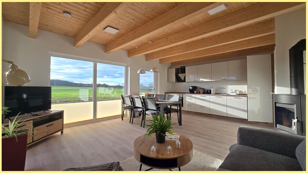 The open living area features a modern kitchen and dining space, with an inviting seating area highlighted by wooden beams overhead. Large windows offer expansive views of the surrounding landscape, while a television is positioned on a sleek cabinet, adding to the space’s functionality.