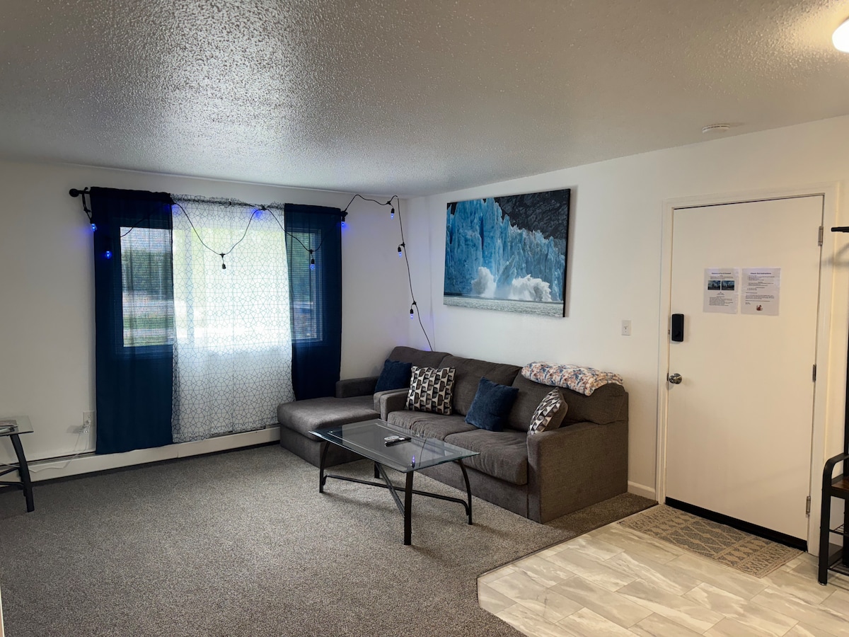 A spacious living area is highlighted by a comfortable gray sofa adorned with decorative pillows. A glass coffee table sits in the center, and a striking piece of art featuring icy blue tones is displayed on the wall. Natural light filters through the window covered by patterned curtains.