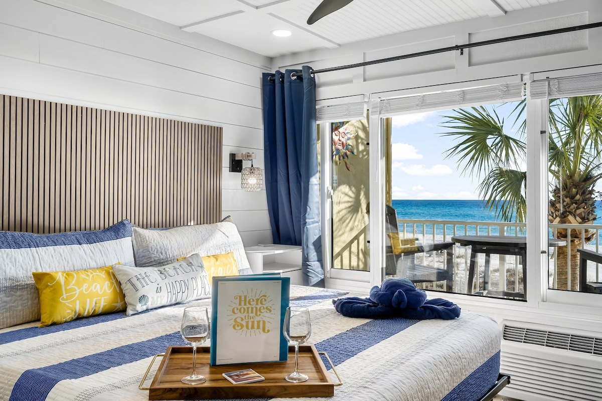 A beach-themed bedroom features a spacious bed adorned with blue and white linens. Large windows provide natural light and views of palm trees and the ocean. A tray with two glasses and a book titled 'Here Comes the Sun' rests on the bed, adding to the inviting decor.