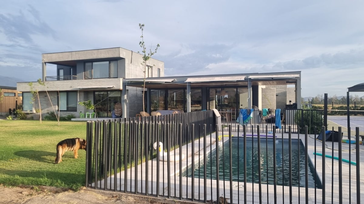 A contemporary two-story home is set within a spacious green area, featuring large glass windows and a fenced pool. A dog stands nearby on the lawn. Loungers and colorful toys are visible around the pool, enhancing the outdoor relaxation space.