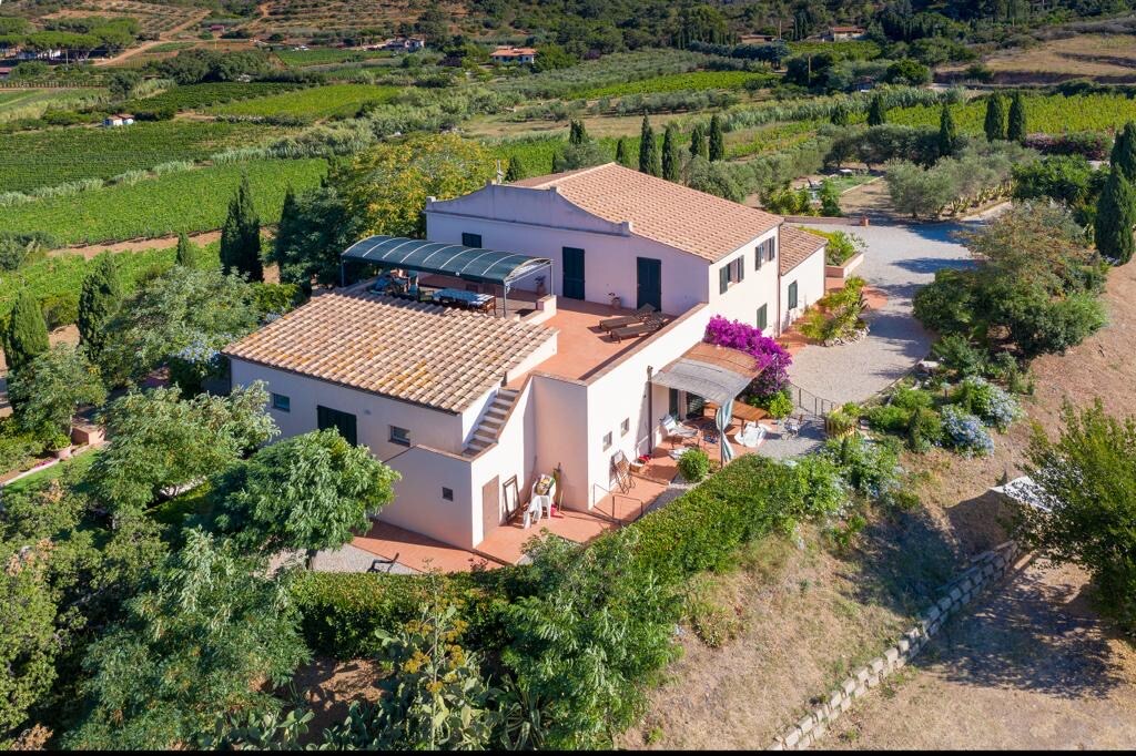 An aerial view of a renovated Tuscan farmhouse is displayed, featuring terracotta roofs and a spacious terrace adorned with flowering plants. Surrounding hills are visible, with a blend of greenery and cultivated land, creating a serene rural landscape.