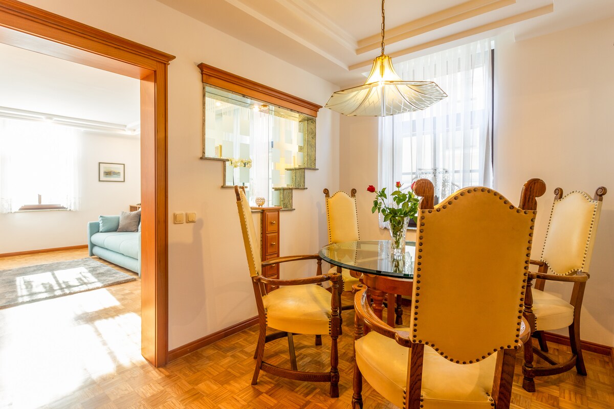 A dining area is presented, featuring a round glass-top table surrounded by six wooden chairs with upholstered backs. Natural light enters through large windows, enhancing the space. A sideboard and decorative glass shelving are visible, complementing the warm wood tones of the room.