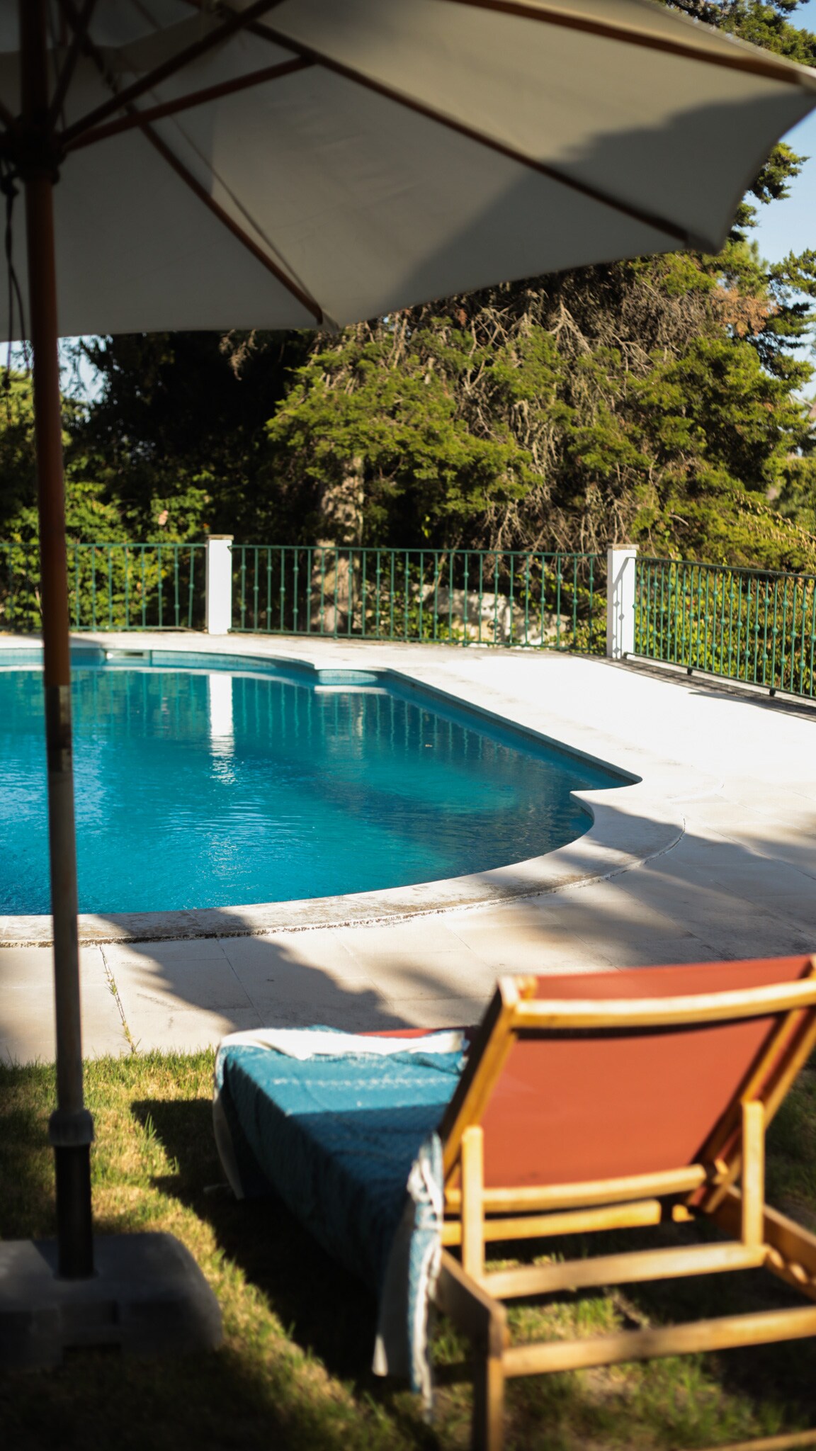 A refreshing pool area is surrounded by lush greenery, providing a serene atmosphere. A sun lounger is positioned under a large umbrella, inviting relaxation by the water's edge. The clear pool reflects natural light, enhancing the calm and tranquil setting.