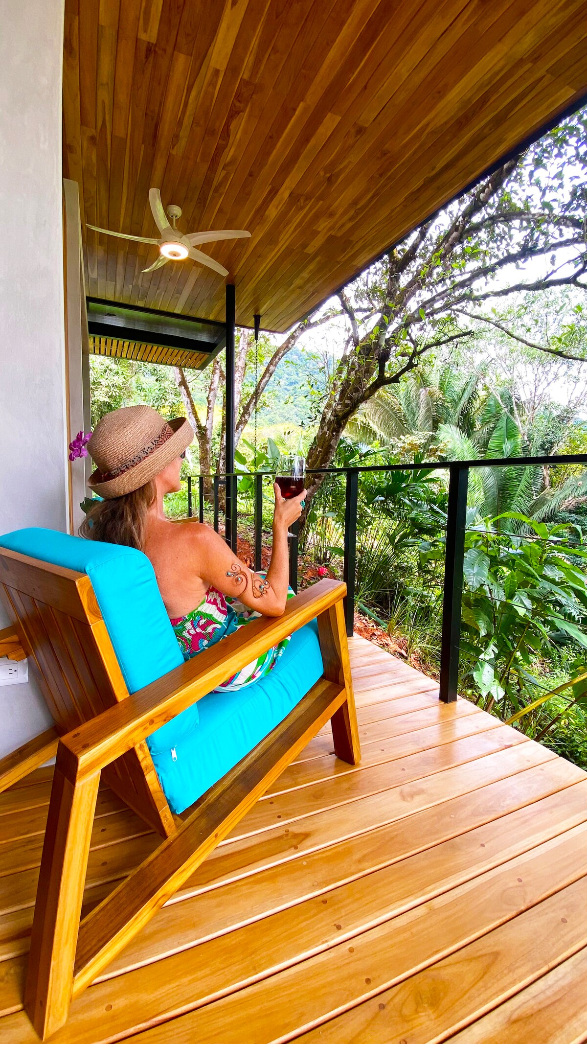 A wooden terrace features a seating area with a bright blue armchair positioned for a scenic view of the dense jungle. A ceiling fan is visible above, and lush greenery surrounds the space, offering a serene and inviting outdoor atmosphere.