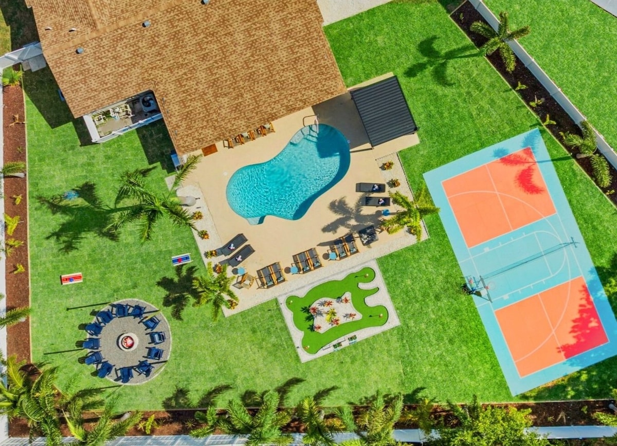 An aerial view showcases a spacious backyard featuring a heated pool with loungers set around it. Nearby, a vibrant basketball and pickleball court occupies part of the neatly manicured lawn, while a fire pit with seating is positioned in a circular pattern, attracting gatherings.