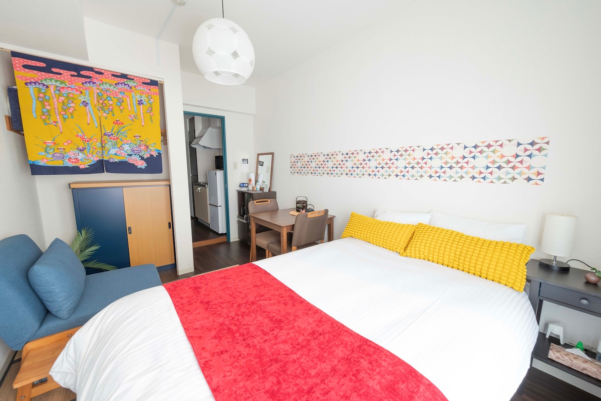 A well-lit room features a semi-double bed with a bright red and white blanket. A small dining area with a table and chairs is visible. Colorful wall art adds a touch of vibrancy to the space, complemented by a sofa in a cozy blue hue.