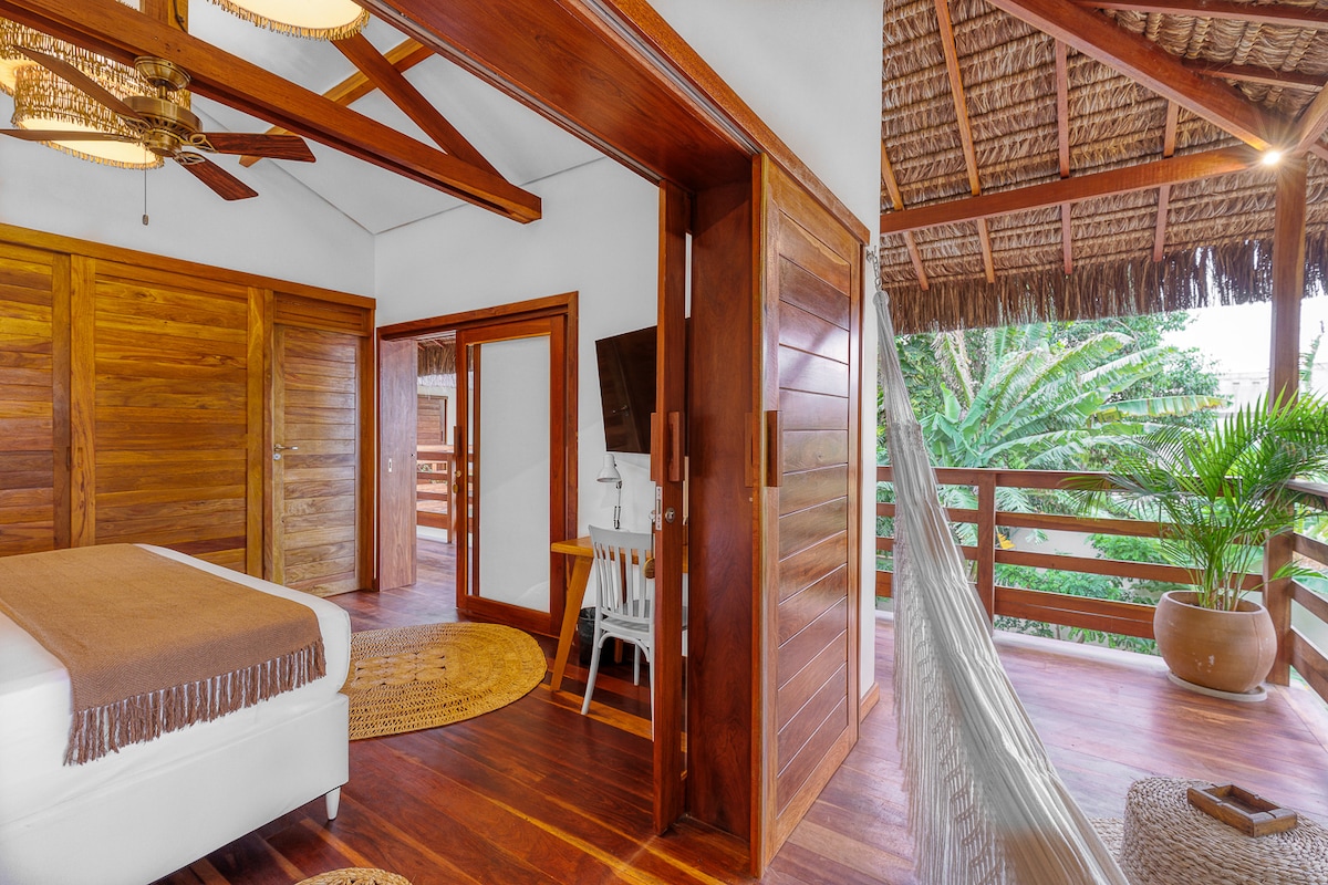 A spacious bedroom features wooden beams and tropical decor, with a large bed draped in a neutral blanket. Access to a private balcony is provided through double doors, allowing natural light to fill the room. A cozy seating area includes a white chair and small table, enhancing comfort.