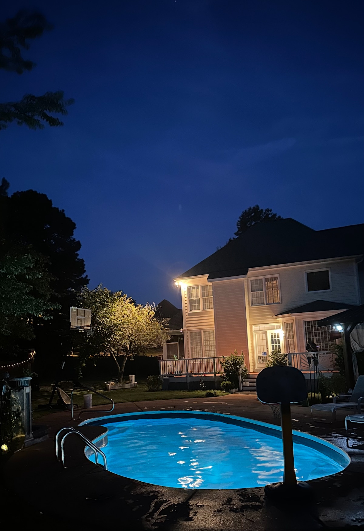A tranquil backyard scene is illuminated by soft lighting, featuring a glowing saltwater pool. The two-story home stands nearby, complemented by a basketball hoop and lush trees. A peaceful evening atmosphere is created by the dimming sky.