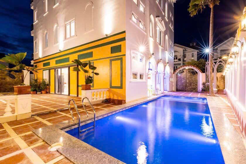 A stylish outdoor area features a large swimming pool with bright blue water, framed by elegant stone tiles. The vibrant yellow and green accents of the building add contrast against the night sky, while ambient lighting enhances the inviting atmosphere.